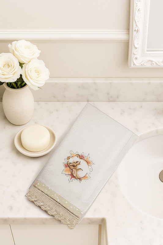 Asciugamano in cotone con ricamo orsetto coccolone addormentato tra foglie e fiori autunnali, fotografato dall’alto su piano bagno in marmo.