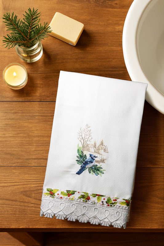Asciugamano Songs of Winter con bluebird ricamato e bordo in pizzo, piegato su piano in legno con candela accesa, sapone e abete in vaso.