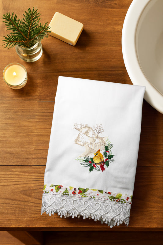 Asciugamano Songs of Winter con cardinale femmina ricamato e bordo natalizio, piegato su piano di legno con candela, sapone e ramoscello di abete.