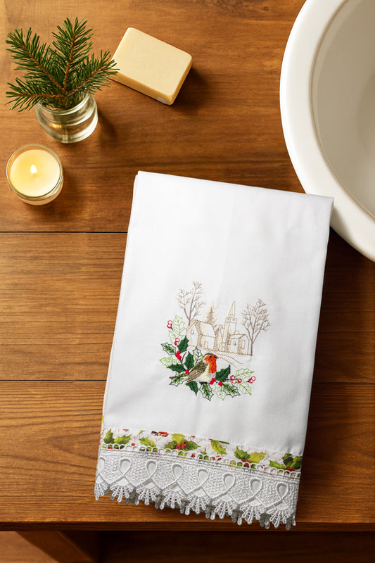 Asciugamano bianco Songs of Winter con pettirosso ricamato e bordo in pizzo, piegato su piano in legno con candela e rametto di abete.