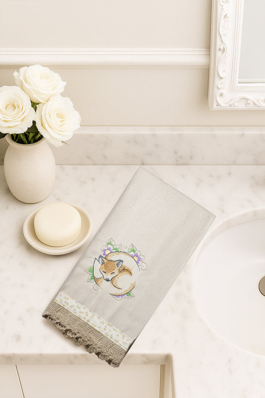 Asciugamano in cotone con ricamo volpe addormentata tra fiori viola e verdi, fotografato dall’alto su piano bagno in marmo.