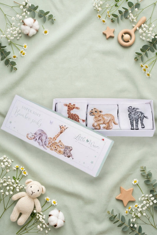 Scatolina regalo Wrendale Designs per neonati decorata con animali della savana: giraffa, elefantino e zebra, contenente tre paia di calzini ricamati, posizionata su fondo verde salvia primaverile con foglie, fiori e giochini.
