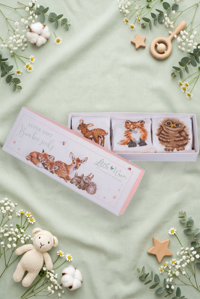 Scatolina regalo Wrendale Designs per neonati con illustrazioni di cerbiatto, volpe e gufetto, contenente tre paia di calzini bianchi con ricami coordinati, posizionata su fondo verde salvia primaverile con foglie, fiori e giochini.