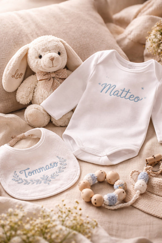 Body bianco personalizzato in vinile, accompagnato da bavaglino ricamato con nome, pupazzo con fiocco e nome ricamato, su fondo neutro, che evocano cura e affetto