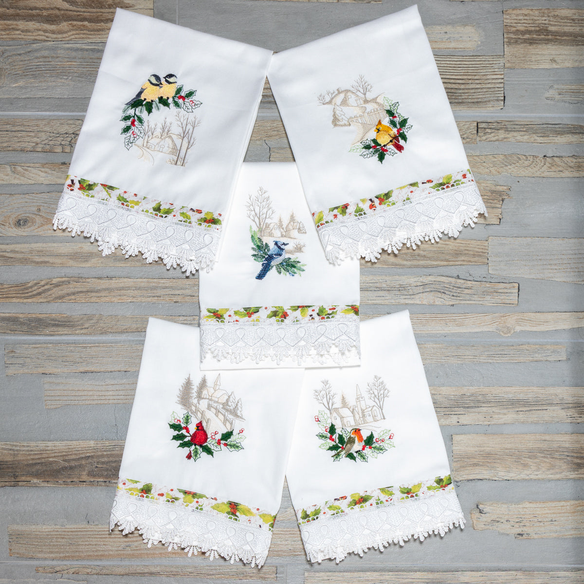 Collezione di asciugamani in cotone bianco Songs of Winter con ricami invernali di paesaggi e uccellini, disposti a ventaglio su fondo legno.
