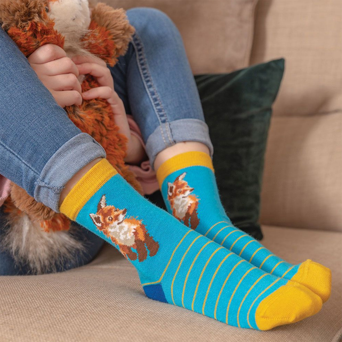 Calze Wrendale Designs da bambino in azzurro e giallo con ricamo di una volpe, indossate mentre il bambino abbraccia un peluche sul divano.