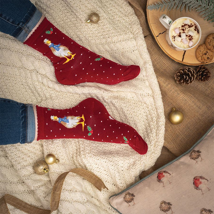 Calze natalizie Wrendale Designs da donna in rosso con ricamo di oca e piccoli dettagli festivi, fotografate su una coperta intrecciata accanto a tazza di cioccolata calda e decorazioni natalizie.