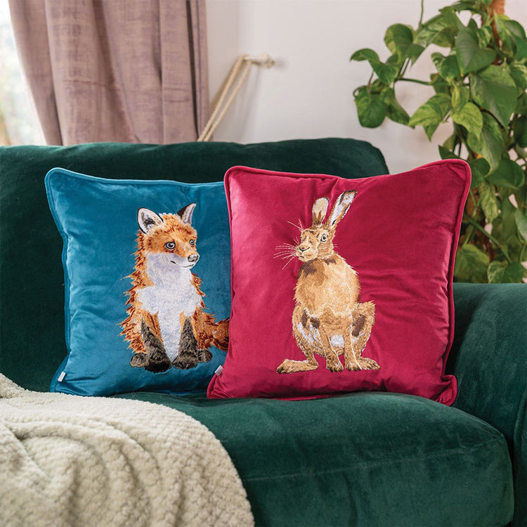 Cuscini Wrendale Designs in velluto, uno blu con ricamo di una volpe e uno rosso con ricamo di una lepre, appoggiati su un divano verde in un salotto accogliente.
