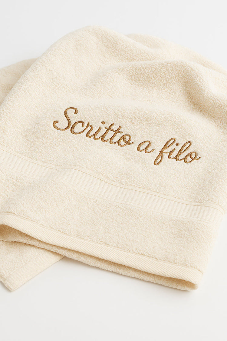Su sfondo neutro, asciugamano in spugna ricamato con la frase Scritto a filo, rappresentativa della collezione di asciugamani personalizzati con nome o iniziali ricamate