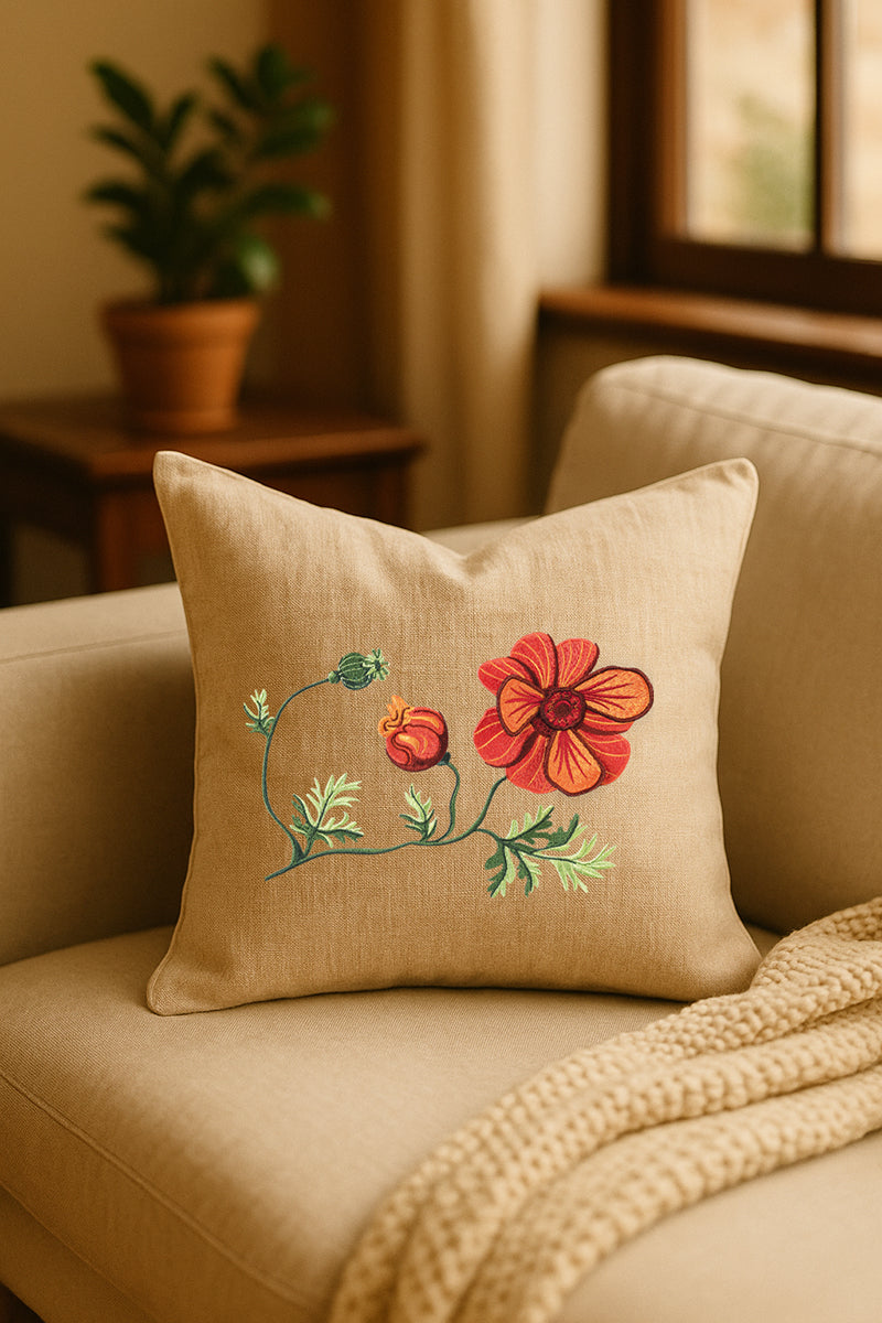 Cuscino in lino beige con ricamo a rilievo di anemone rosso e bocciolo arancione, appoggiato su un divano chiaro in un ambiente caldo e naturale, con pianta verde sullo sfondo.