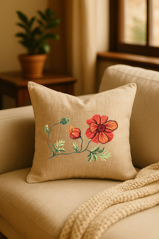 Cuscino in lino beige con ricamo a rilievo di anemone rosso e bocciolo arancione, appoggiato su un divano chiaro in un ambiente caldo e naturale, con pianta verde sullo sfondo.
