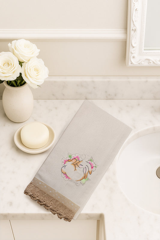 Asciugamano in cotone con ricamo coniglietto sognatore circondato da fiori rosa e verdi, fotografato dall’alto su piano bagno in marmo.