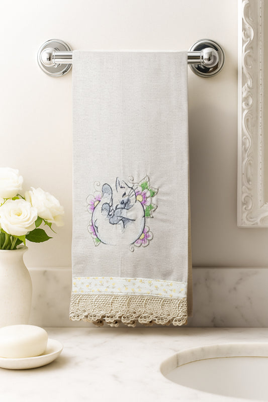 Asciugamano in cotone con ricamo gattino dei fiori arrotolato tra fiori lilla e verdi, fotografato appeso in un bagno elegante.