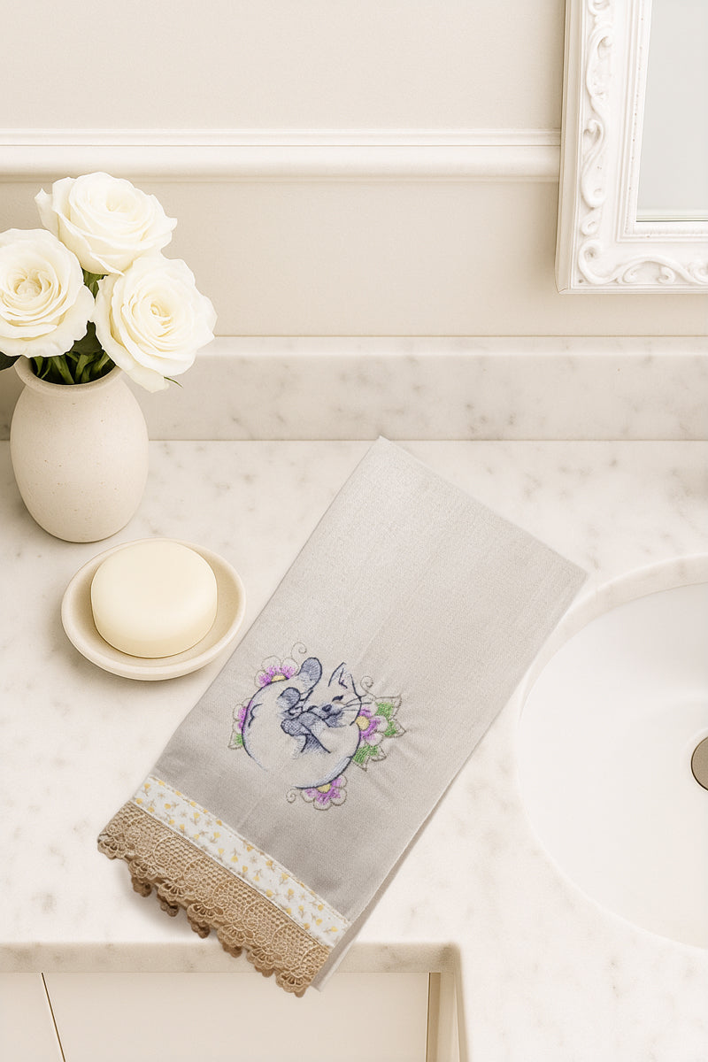 Asciugamano in cotone con ricamo gattino dei fiori arrotolato tra fiori lilla e verdi, fotografato dall’alto su piano bagno in marmo.