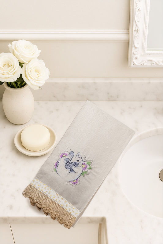 Asciugamano in cotone con ricamo gattino dei fiori arrotolato tra fiori lilla e verdi, fotografato dall’alto su piano bagno in marmo.