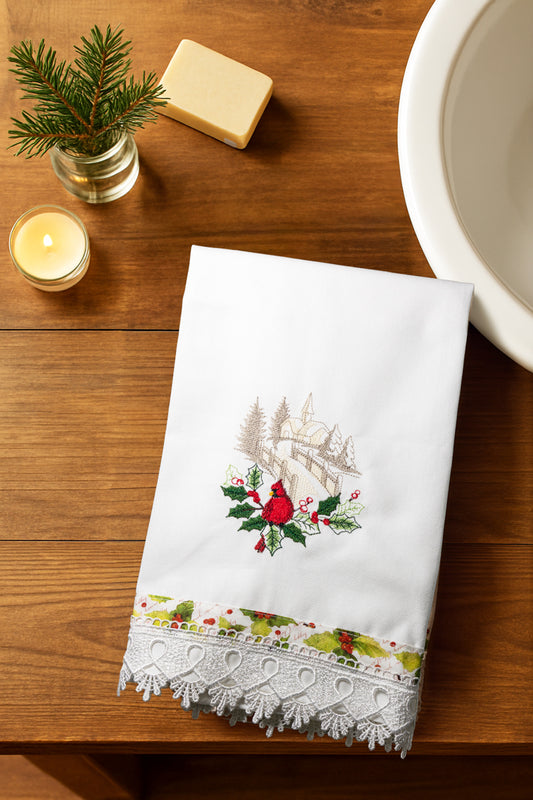 Asciugamano Songs of Winter con cardinale rosso ricamato e bordo in pizzo, piegato su piano in legno con candela, sapone e decorazioni natalizie.