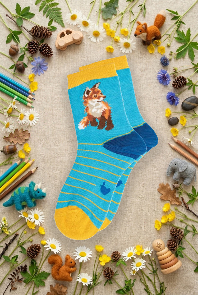 Calze Wrendale Designs da bambini in azzurro e giallo con volpe ricamata, presentate su sfondo naturale primaverile, animaletti in feltro e matite colorate.
