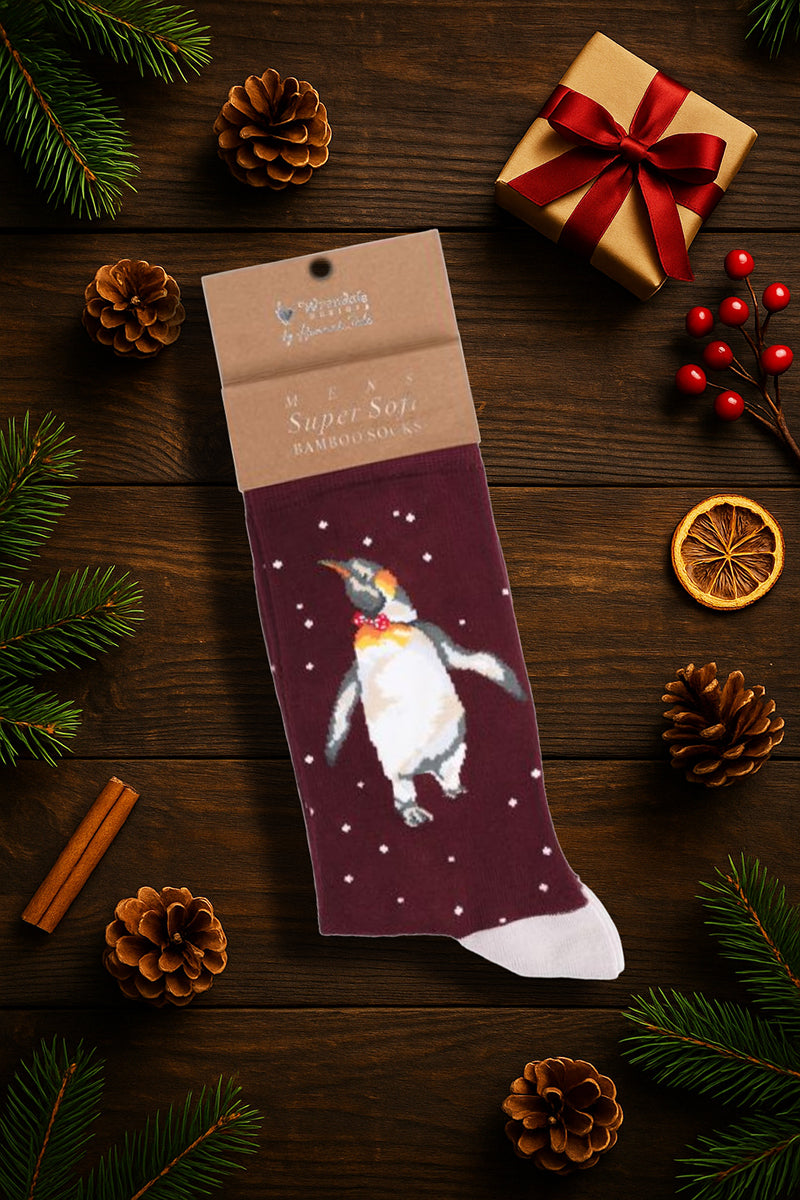 Calza da uomo Wrendale Designs in bambù bordeaux con pinguino ricamato con papillon rosso, fotografata in ambientazione natalizia con pigne, rami di abete e decorazioni festive.