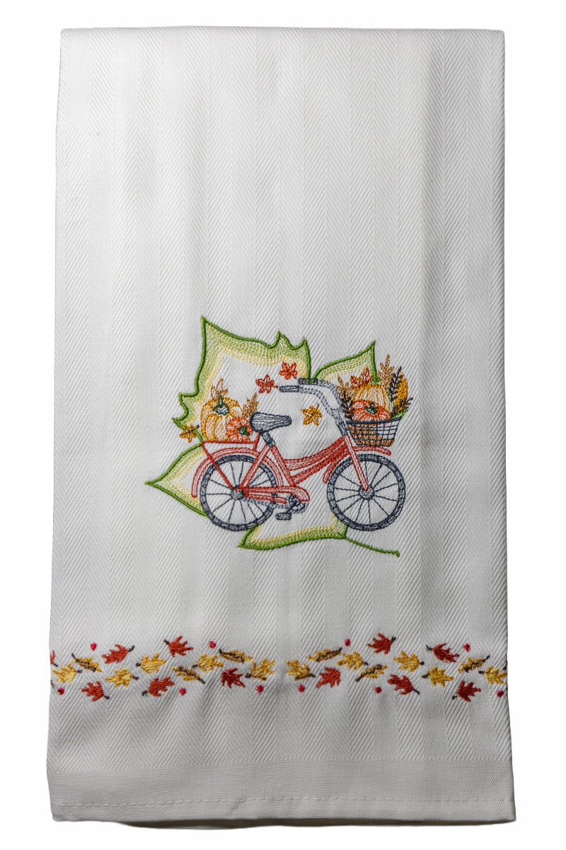 Ricamo bicicletta con cestino autunnale e foglie colorate su asciugapiatto in cotone bianco, fotografato su sfondo neutro.