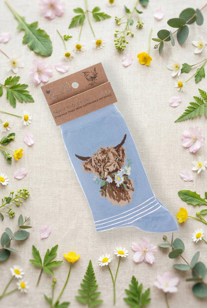 Calza da donna Wrendale Designs in bambù azzurro con mucca delle Highland e margherite ricamate, fotografata su sfondo primaverile con fiori e foglie.