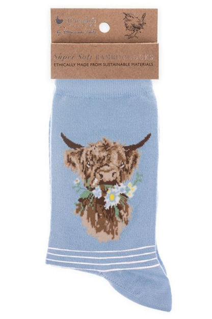 Calza da donna Wrendale Designs in bambù azzurro con ricamo di mucca Highland e mazzolino di margherite, presentata con cartoncino kraft del brand su sfondo neutro.