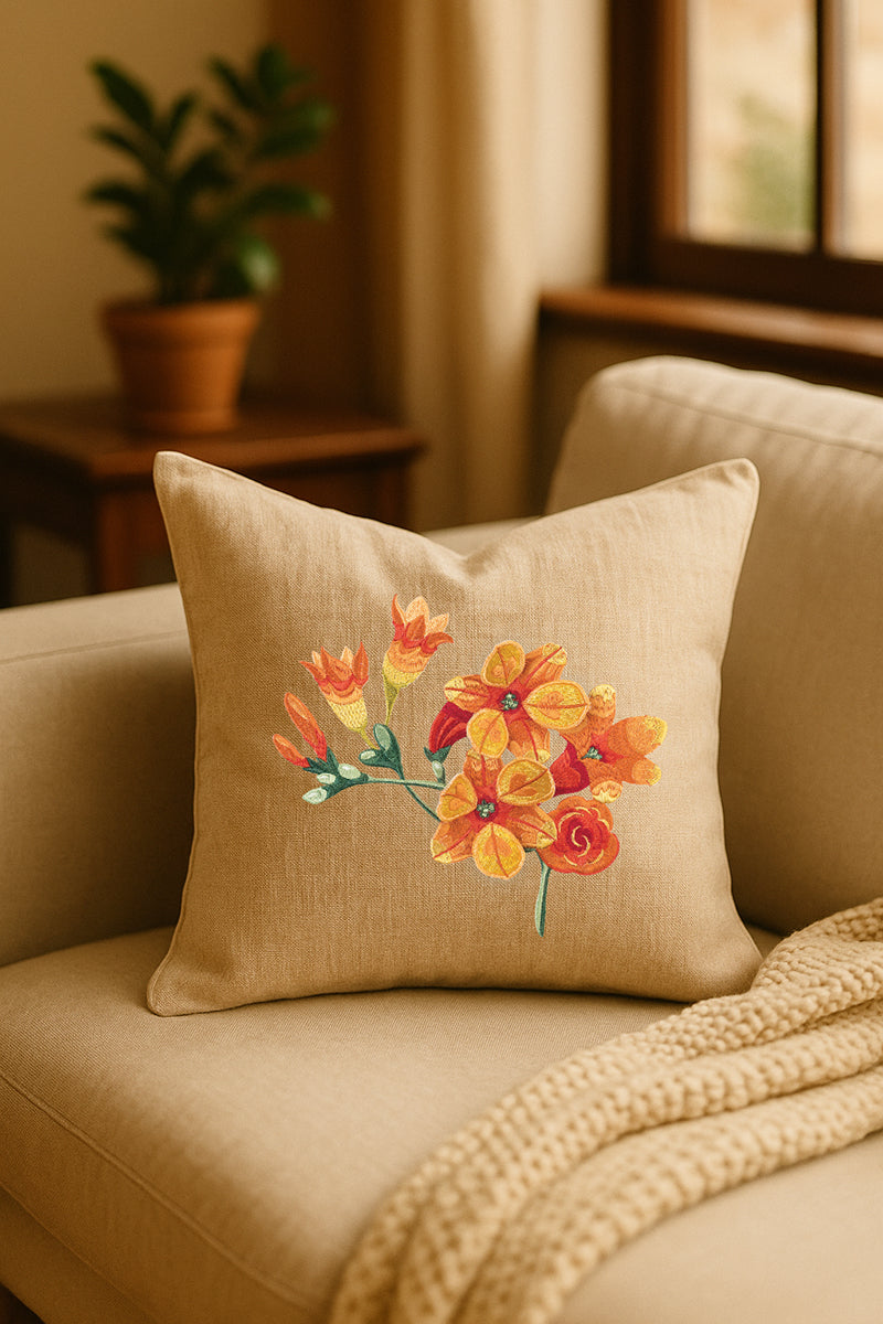 Cuscino decorativo in misto lino beige con ricamo a rilievo di fresie arancioni e gialle, appoggiato su un divano chiaro in un ambiente caldo e naturale con pianta sullo sfondo.