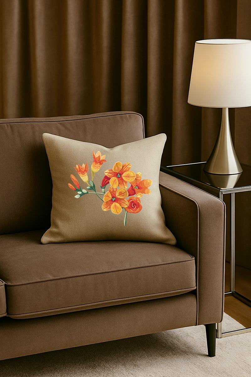 Cuscino in misto lino beige con ricamo 3D di fresie arancioni, posato su un divano marrone in un soggiorno elegante con lampada dorata e tende scure.