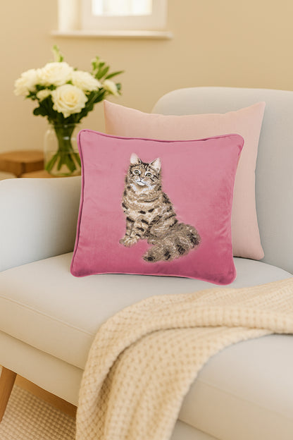Cuscino in velluto rosa Wrendale Designs con ricamo di un gatto tigrato, posato su un divano chiaro in un salotto con fiori sullo sfondo.