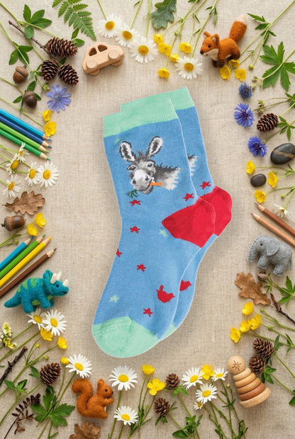 Calze Wrendale Designs da bambini in azzurro e rosso con asinello ricamato, presentate su sfondo naturale primaverile, animaletti in feltro e matite colorate.