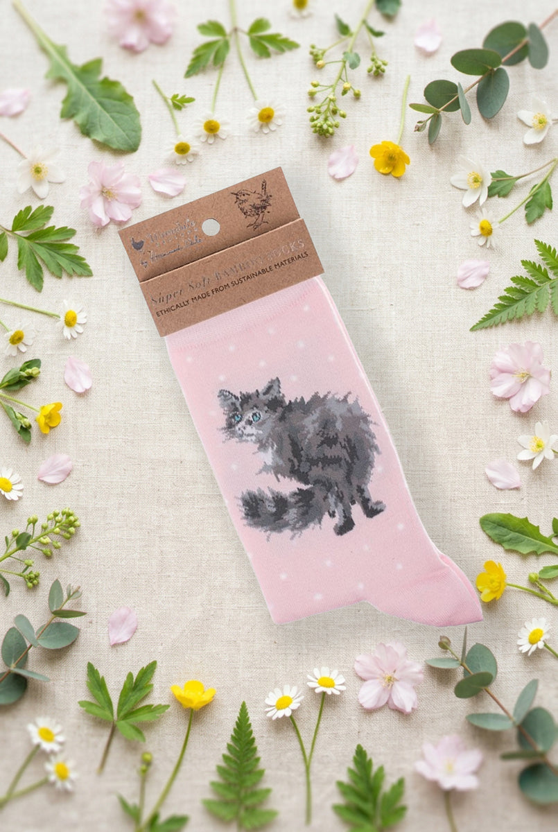 Calza da donna Wrendale Designs in bambù rosa chiaro con gatto soffice grigio ricamato e piccoli pois bianchi, fotografata su sfondo primaverile con fiori e foglie.