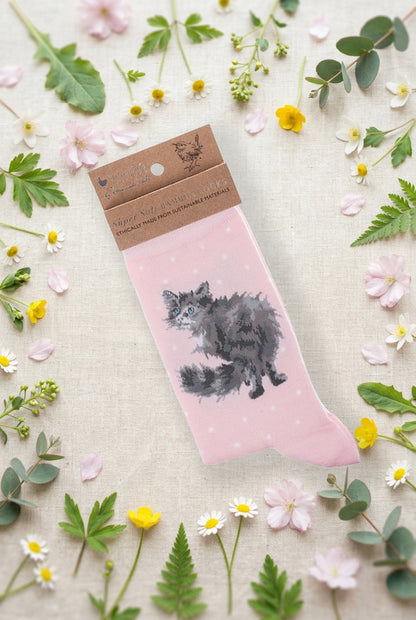 Calza da donna Wrendale Designs in bambù rosa chiaro con gatto soffice grigio ricamato e piccoli pois bianchi, fotografata su sfondo primaverile con fiori e foglie.