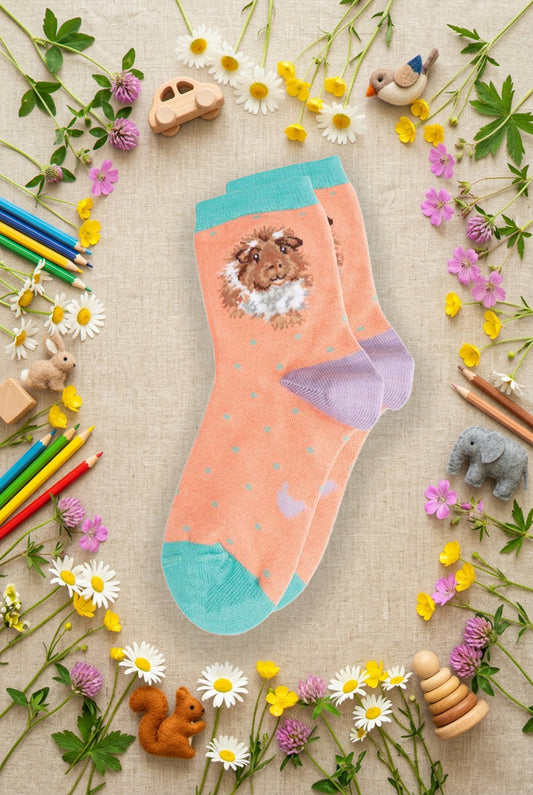 Calze Wrendale Designs da bambini in rosa con porcellino d'India ricamato, presentate su sfondo naturale primaverile, animaletti in feltro e matite colorate.