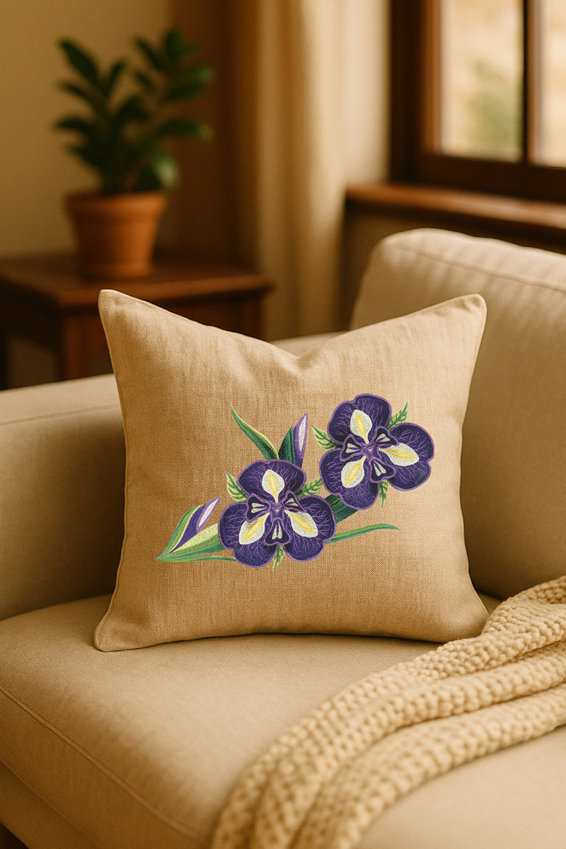 Cuscino decorativo in misto lino beige con ricamo tridimensionale di iris viola, appoggiato su un divano chiaro in un ambiente caldo e naturale con pianta sullo sfondo.