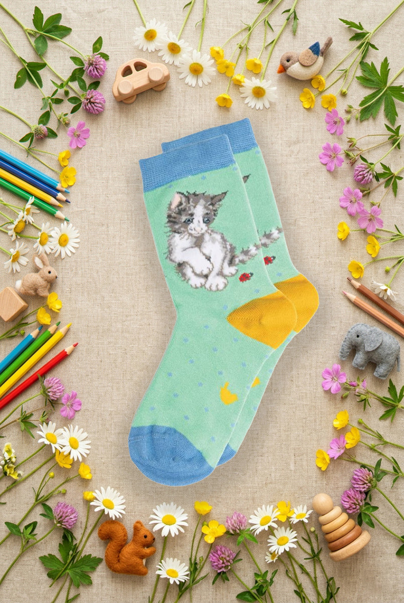 Calze Wrendale Designs da bambini in verde acqua e giallo con gattino e coccinella ricamate, presentate su sfondo naturale primaverile, animaletti in feltro e matite colorate.