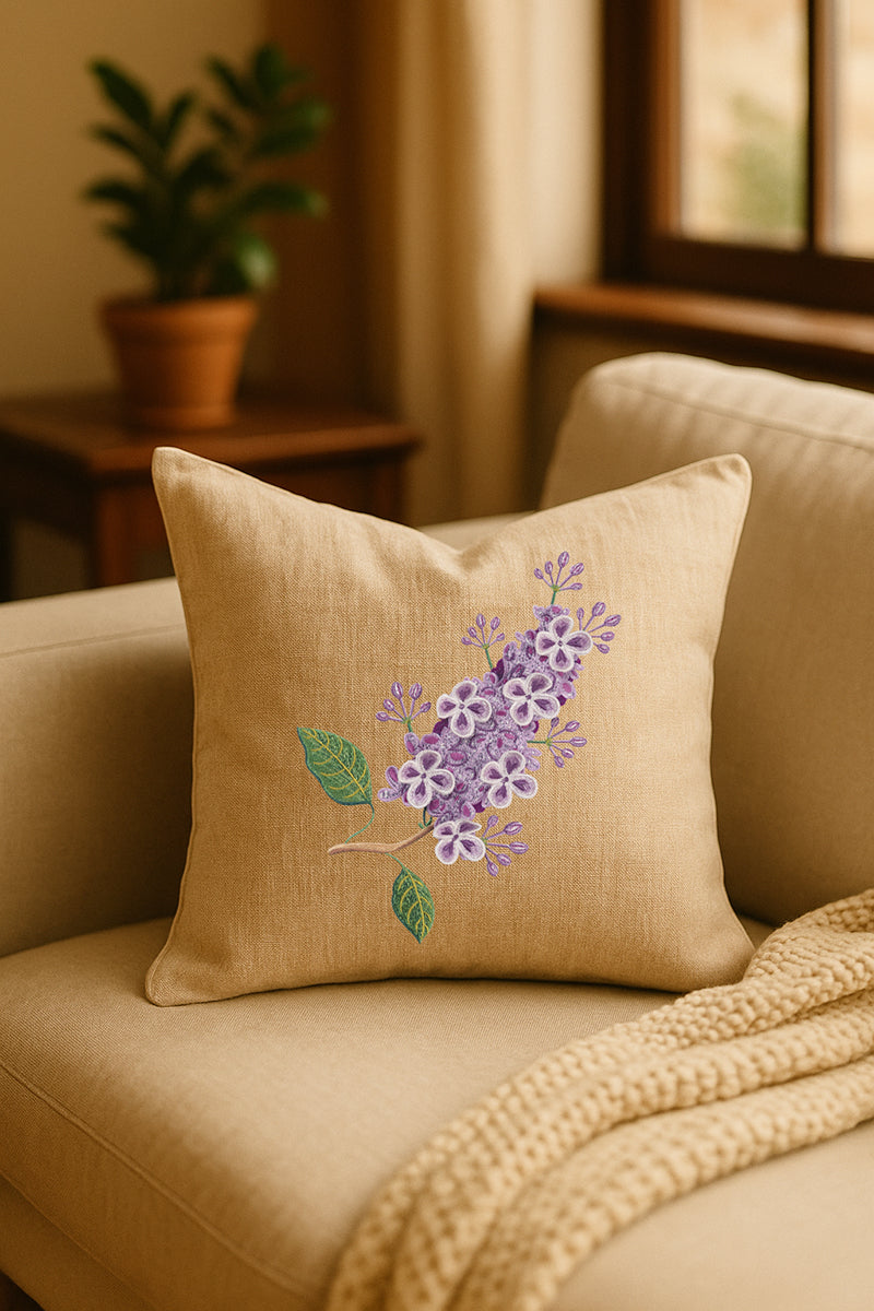 Cuscino decorativo in misto lino beige con ricamo tridimensionale di lillà lilla, posato su un divano chiaro in un ambiente naturale e luminoso con pianta sullo sfondo.