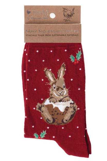 Calza da donna Wrendale Designs in bambù rosso con ricamo di coniglio natalizio seduto su un pudding decorato e piccoli pois bianchi, presentata con cartoncino kraft del brand su sfondo neutro.
