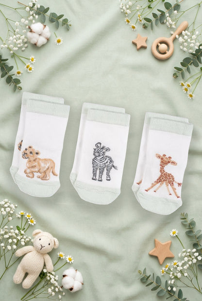 Tre paia di calzini da neonato Wrendale Designs in bianco con polsino verde chiaro, ciascuno con ricamo di giraffina, zebra o leoncino, mostrati su sfondo verde salvia primaverile con foglie, fiori e giochini.