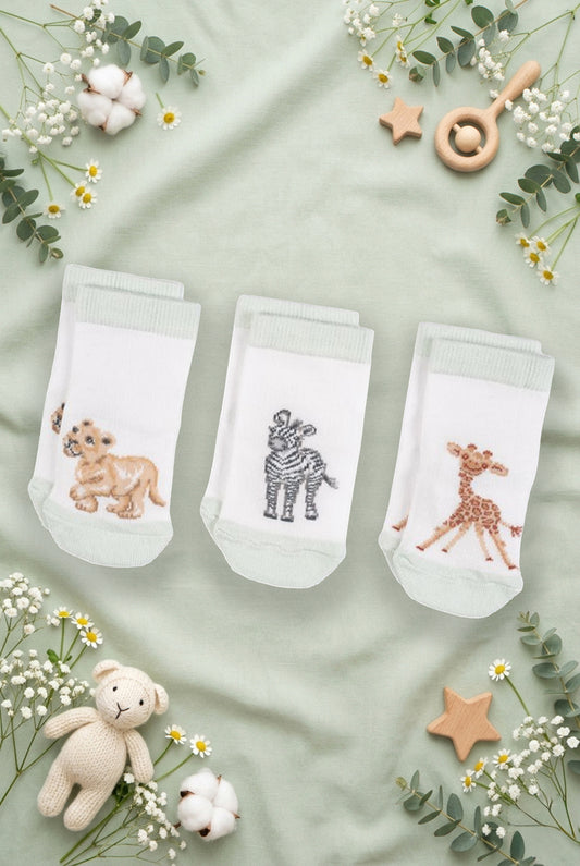 Tre paia di calzini da neonato Wrendale Designs in bianco con polsino verde chiaro, ciascuno con ricamo di giraffina, zebra o leoncino, mostrati su sfondo verde salvia primaverile con foglie, fiori e giochini.