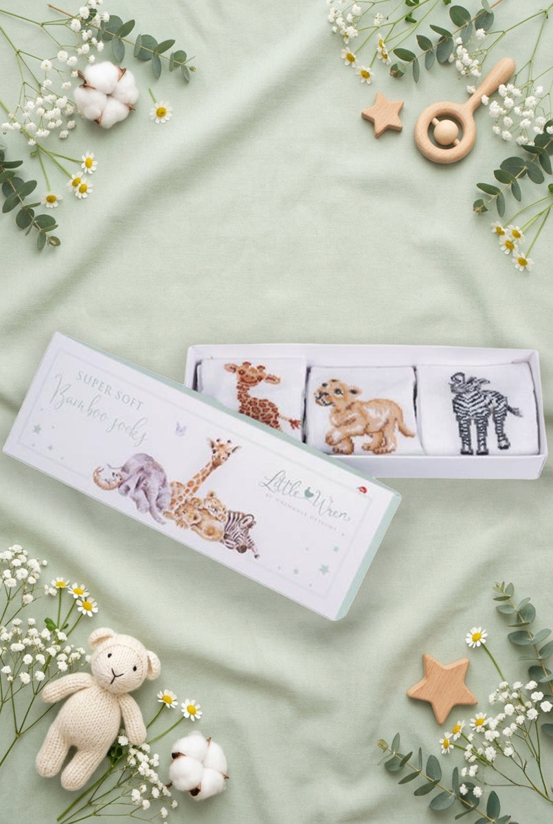 Scatolina regalo Wrendale Designs per neonati decorata con animali della savana: giraffa, elefantino e zebra, contenente tre paia di calzini ricamati, posizionata su fondo verde salvia primaverile con foglie, fiori e giochini.