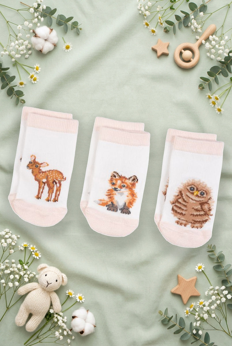 Tre paia di calzini da neonato Wrendale Designs in bianco con polsino rosa, ciascuno decorato con ricamo di cerbiatto, volpe o gufetto, mostrati su sfondo verde salvia primaverile con foglie, fiori e giochini.
