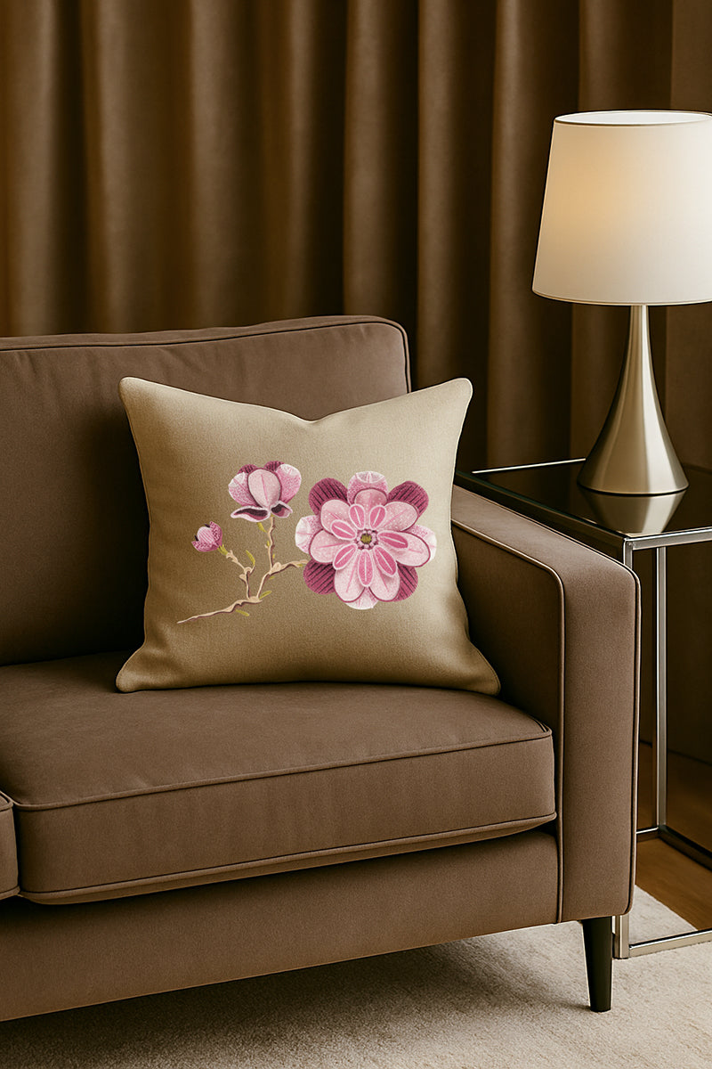 Cuscino decorativo in misto lino beige caldo con ricamo tridimensionale di magnolia rosa, posato su un divano marrone in un soggiorno elegante con lampada dorata e tende scure.