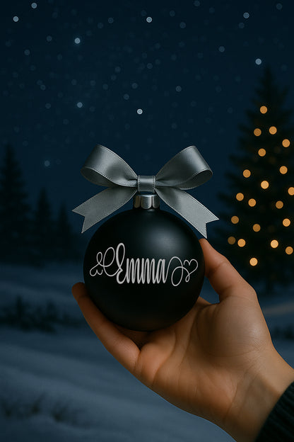 Pallina di Natale in vetro nero con fiocco elegante e personalizzazione in vinile color argento, fotografata su esterno invernale con stelle e luci soffuse.