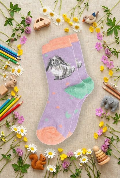 Calze Wrendale Designs da bambini in lilla e verde acqua con coniglietto ricamato, presentate su sfondo naturale primaverile, animaletti in feltro e matite colorate.