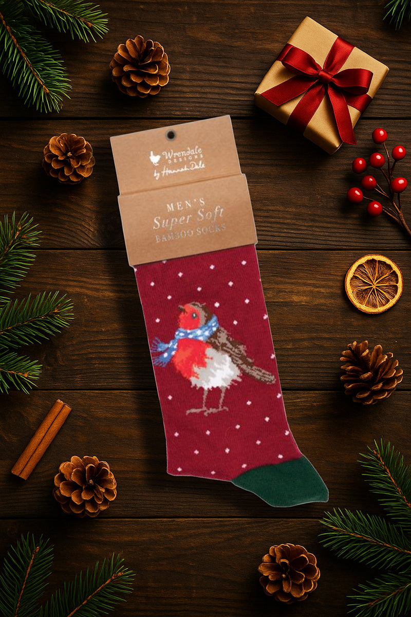 Calza da uomo Wrendale Designs in bambù rosso con illustrazione di un pettirosso con sciarpa, fotografata in ambientazione natalizia su sfondo in legno con pigne, bacche rosse e pacchetto regalo.