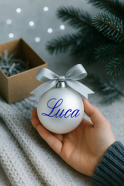 Pallina di Natale in vetro bianco perlaceo con fiocco elegante e personalizzazione in vinile blu, fotografata su fondo festivo con luci soffuse.